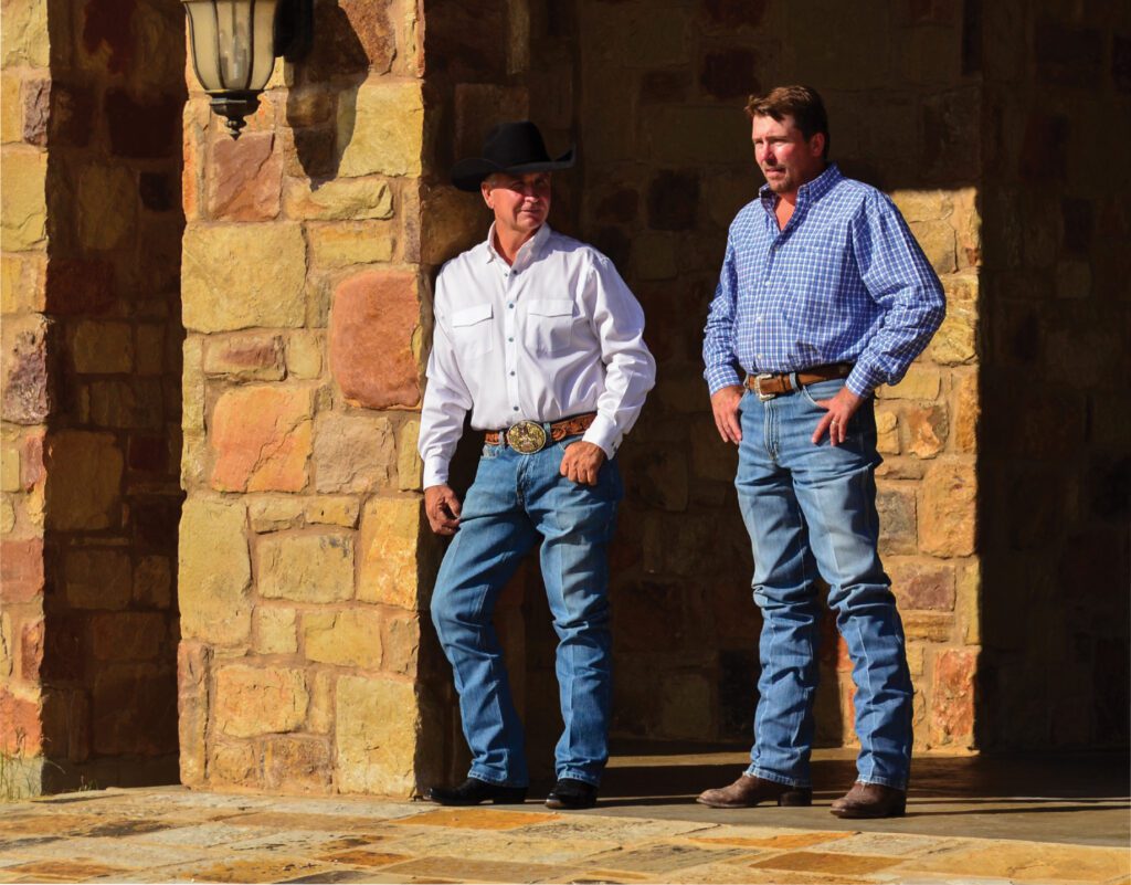 Two cowboys in western attire, standing against a stone wall, embodying rustic lifestyle and culture. Sunlight highlights their casual stance.