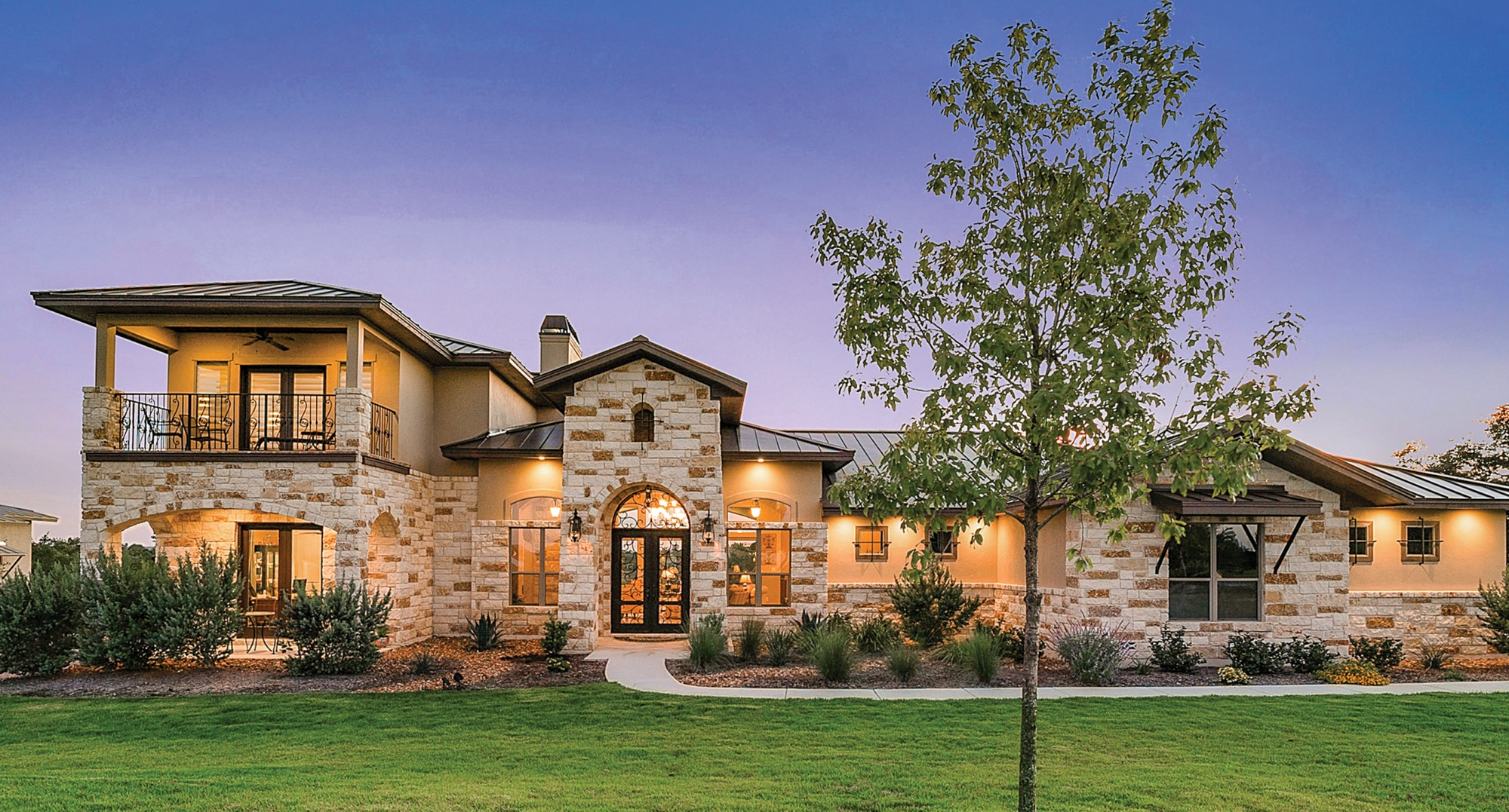 Modern stone house with an inviting entrance, balcony, and landscaped yard, highlighted by soft evening light against a twilight sky.