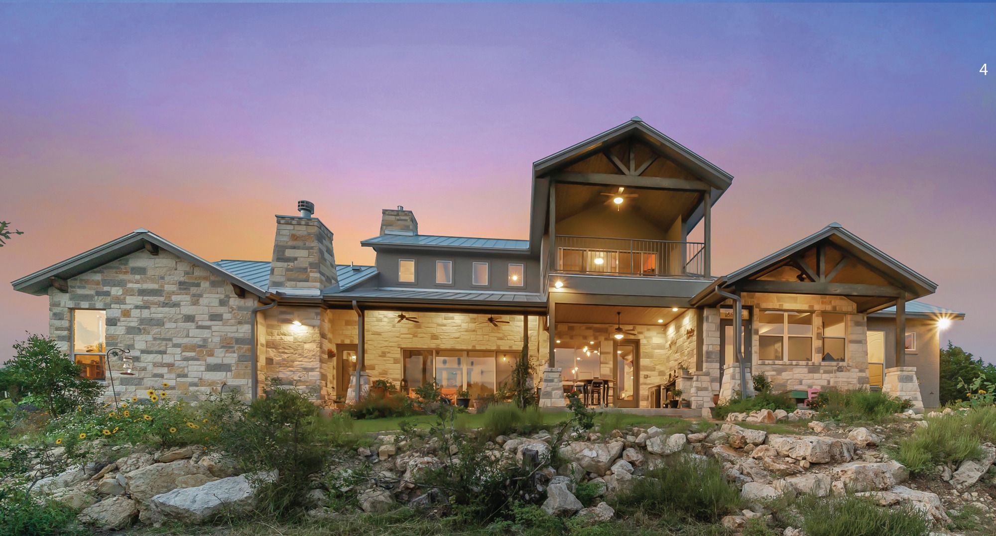 Modern stone house with large windows and a balcony, set against a twilight sky. Lush greenery and rocky landscape surround the structure, emphasizing its design.