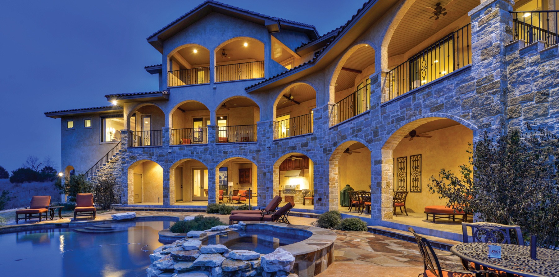 Elegant multi-level home with stone and stucco exterior, featuring arched balconies and a poolside setup, illuminated at dusk.